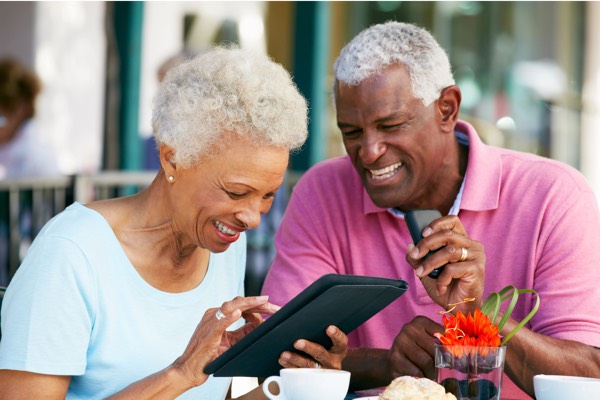 Smiling senior couple at an outdoor café table, joyfully looking at a tablet together, with coffee cups, a pastry, and a vibrant flower centerpiece in the foreground.